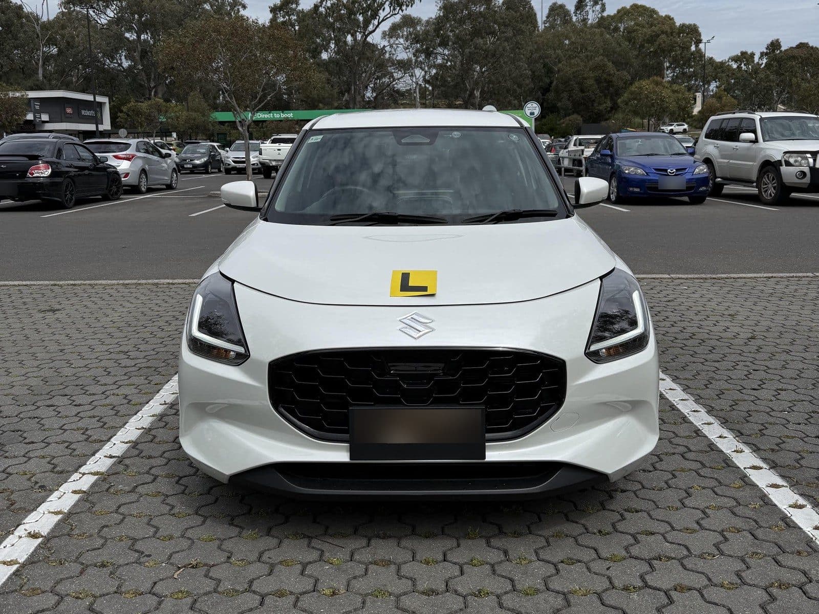 The front of Stacey's white Suzuki Swift learner car, with a yellow L plate on the bonnet